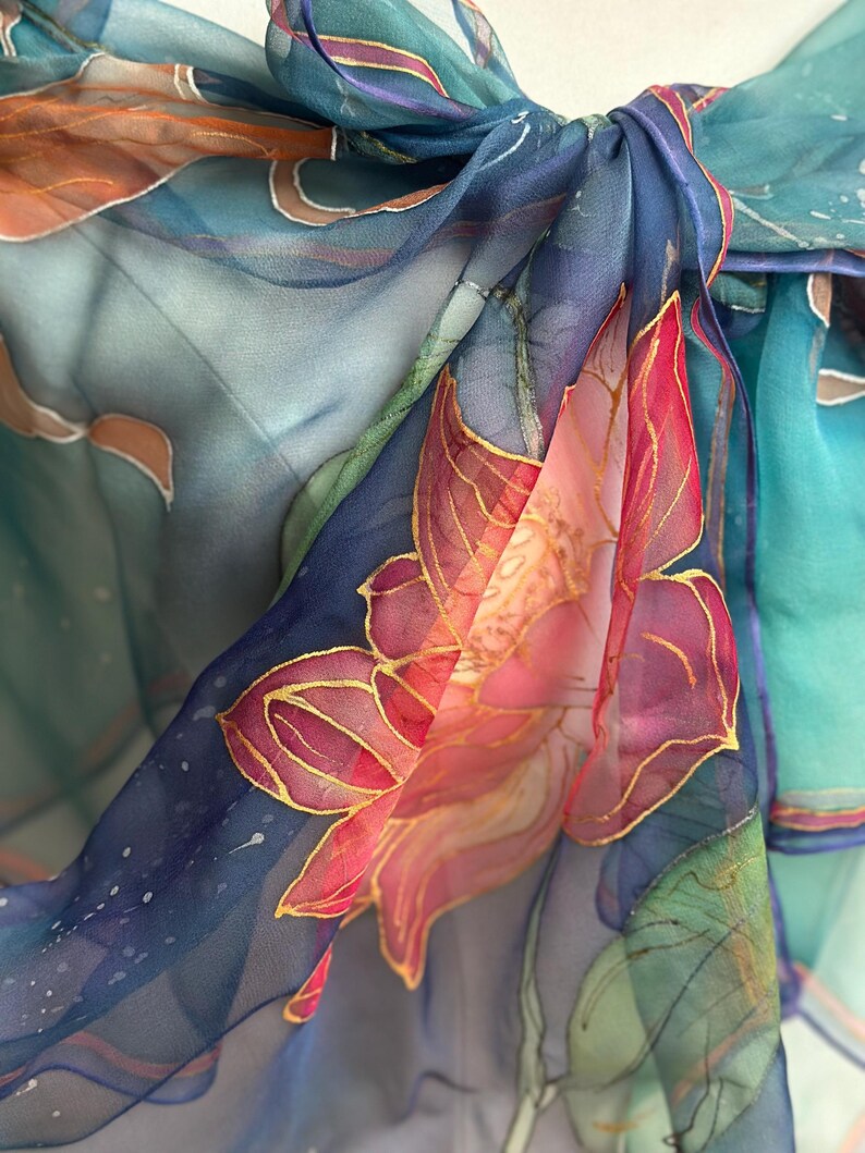 May include: Close-up of a sheer, flowing scarf with a hand-painted floral design. The scarf features a vibrant color palette of blues, pinks, and greens, with gold accents outlining the floral details. The fabric is knotted at the top.