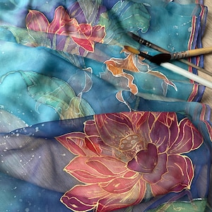 May include: A close-up of a silk scarf with a hand-painted floral design. The scarf features large lotus flowers in shades of pink, red, and gold against a blue background. Paintbrushes are visible, suggesting the artwork is in progress.