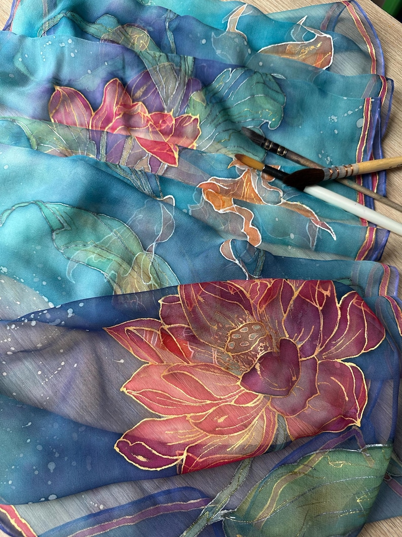 May include: A close-up of a silk scarf with a hand-painted floral design. The scarf features large lotus flowers in shades of pink, red, and gold against a blue background. Paintbrushes are visible, suggesting the artwork is in progress.