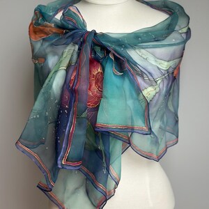May include: A sheer, teal-colored scarf with an abstract design, draped over a mannequin. The scarf features a watercolor-like pattern with hints of orange, purple, and gold accents. The edges are trimmed with a thin gold border.