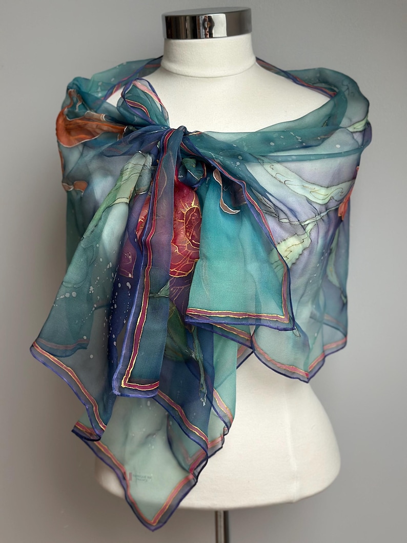 May include: A sheer, teal-colored scarf with an abstract design, draped over a mannequin. The scarf features a watercolor-like pattern with hints of orange, purple, and gold accents. The edges are trimmed with a thin gold border.