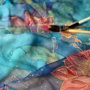 May include: Close-up of a silk scarf with a floral design in shades of blue, teal, and pink. The scarf features hand-painted flowers with gold outlines and a watercolor effect. Paintbrushes are visible in the background.