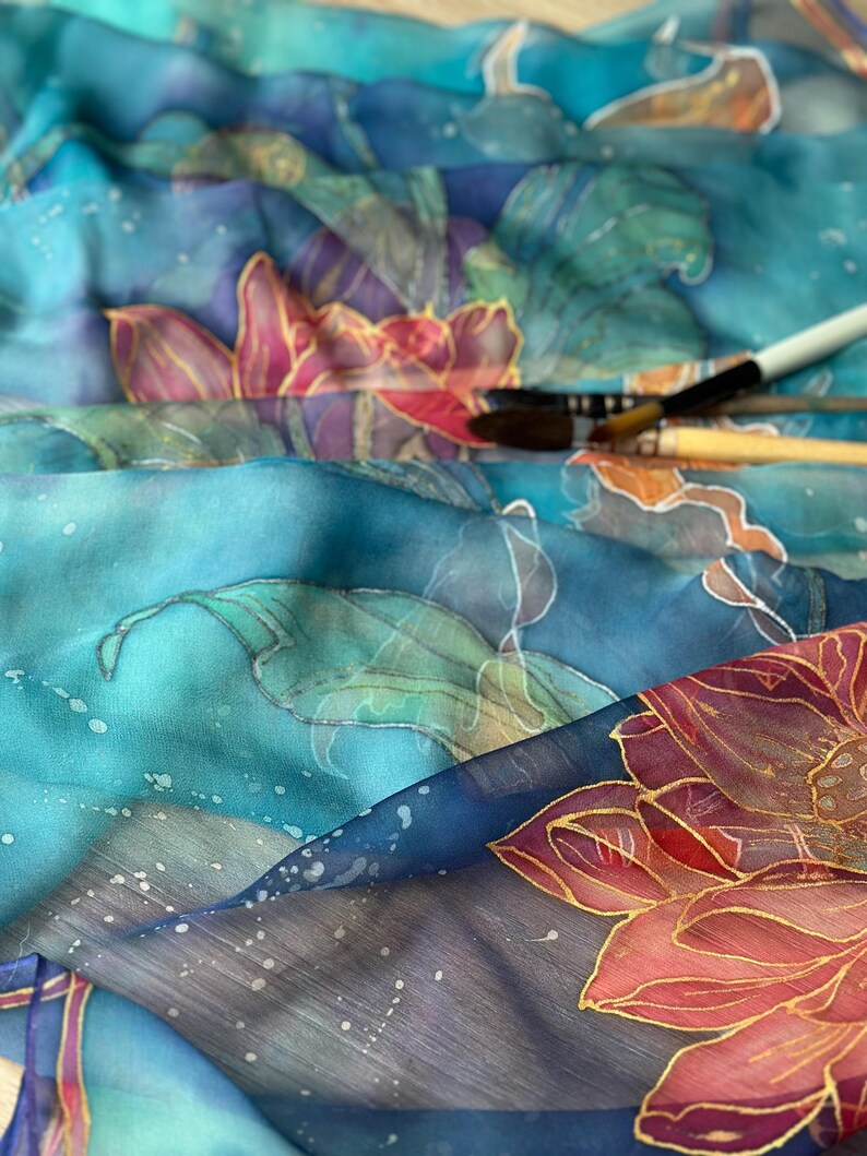 May include: Close-up of a silk scarf with a floral design in shades of blue, teal, and pink. The scarf features hand-painted flowers with gold outlines and a watercolor effect. Paintbrushes are visible in the background.