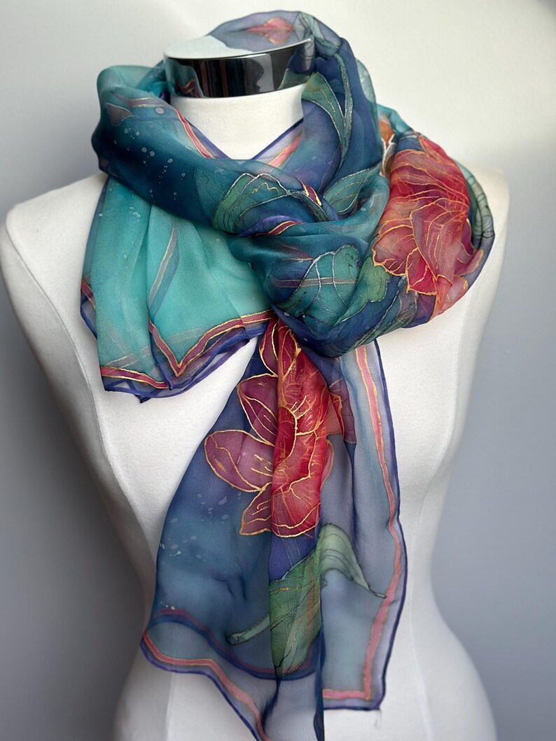 May include: A sheer silk scarf with a floral design. The scarf features a color palette of teal, navy, and red, with gold accents. The scarf is draped over a white mannequin, showcasing its flowing fabric and intricate details.