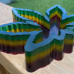May include: A clear glass ashtray shaped like a marijuana leaf. The ashtray is layered with red, yellow, and green resin, creating a colorful and vibrant design.