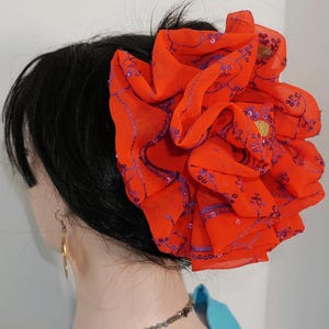 May include: A large, bright orange flower hair clip with purple and gold accents. The flower is made of fabric and has a ruffled texture.