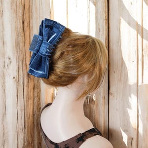 May include: A blue denim bow hair clip with a textured pattern. The clip is worn in a woman's hair.