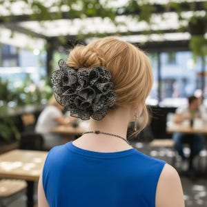 以下が含まれることがあります： 黒と白のレースのヘアクリップは、大きなフリルの花の形をしており、金髪の人の髪に着用されています。 その人は青いトップスを着ています。 背景はぼやけており、屋外環境を示唆しています。