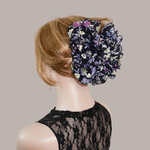 May include: A large, ruffled hair accessory featuring a dark navy blue fabric base with a vibrant floral pattern in shades of purple, yellow, and light blue. The accessory is constructed with multiple layers of fabric, creating a voluminous, circular shape.