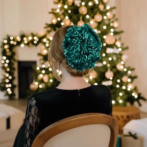 Peut inclure: Un accessoire de cheveux circulaire vert foncé recouvert de petites paillettes réfléchissantes. L'accessoire est froncé en une forme volumineuse et ondulée, placé à l'arrière de la tête sur des cheveux châtain clair coiffés en chignon.