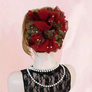 Op de afbeelding: Een bordeauxrood stoffen haaraccessoire met gouden borduursel en parelaccenten. Het accessoire is gestyled op een mannequinhoofd met een zwarte kanten top en een parelketting met dubbele streng. De achtergrond is zachtroze.