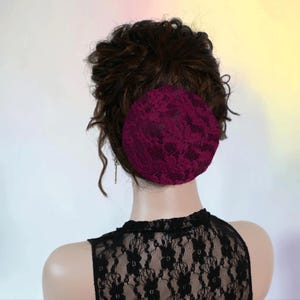 May include: A circular burgundy hair bun cover featuring a detailed black floral lace overlay. The accessory is positioned over dark brown hair styled in a bun, paired with a black lace sleeveless top that has a matching floral pattern.
