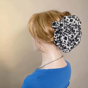 May include: A black and white geometric patterned hair accessory, resembling a flower, is attached to the side of a mannequin's blonde hair. The mannequin is wearing a blue top and a silver necklace. The hair accessory is the focal point.