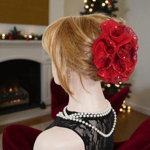 May include: A red sequined flower hair clip is attached to a mannequin's blonde hair. The mannequin wears a black lace top and a white pearl necklace. A fireplace and Christmas tree are visible in the background.