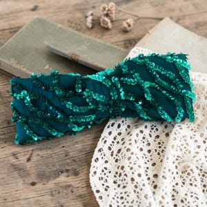 Può includere: Una fascia per capelli in tessuto verde ottanio scuro con un design attorcigliato, decorata con file di paillettes verdi scintillanti abbinate, disposte in un motivo ondulato e organico su tutta la superficie dell'accessorio.