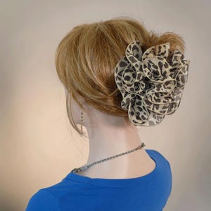以下が含まれることがあります： 黒とタンの色合いのヒョウ柄模様の、大きな花型のヘアアクセサリー。明るい茶色の髪に装着されています。青いシャツとシルバーのネックレスを着用しています。全体的にエレガントなスタイルです。