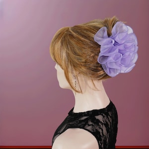 May include: A light purple organza hair scrunchie with a ruffled, layered design. The accessory is worn in a high bun on a mannequin head with light brown hair, styled with side-swept bangs and a black lace top visible at the neckline.