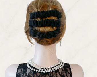 Puede incluir: Un accesorio para el cabello negro con tres bandas elásticas paralelas cubiertas de tela conectadas por peines laterales, sujeto en cabello castaño claro. El accesorio se lleva sobre un collar de perlas blancas de tres hilos y una blusa de encaje negro.