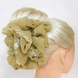 May include: A gold chiffon hair scrunchie with gold and silver sequins and embroidery. The scrunchie is tied in a large bow and is worn in a woman's hair.