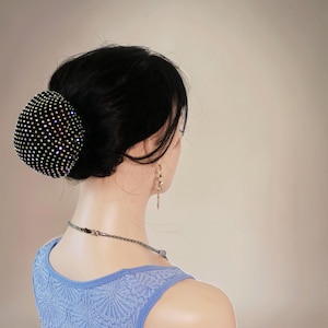 May include: A black hair bun cover embellished with many sparkling rhinestones. The bun cover is round and completely covered in rhinestones, producing a dazzling effect. The model is wearing a light blue top and a silver necklace.
