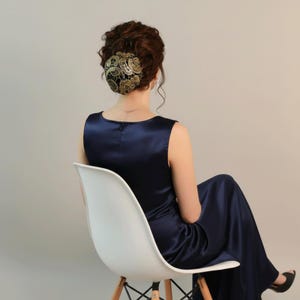 Könnte beinhalten: Ein bodenlanges, ärmelloses Kleid aus marineblauem Satin mit Rundhalsausschnitt. Das Haar ist zu einem Dutt hochgesteckt, der mit einem schwarz-goldenen Spitzen-Haaraccessoire mit detailliertem Blumenmuster bedeckt ist.