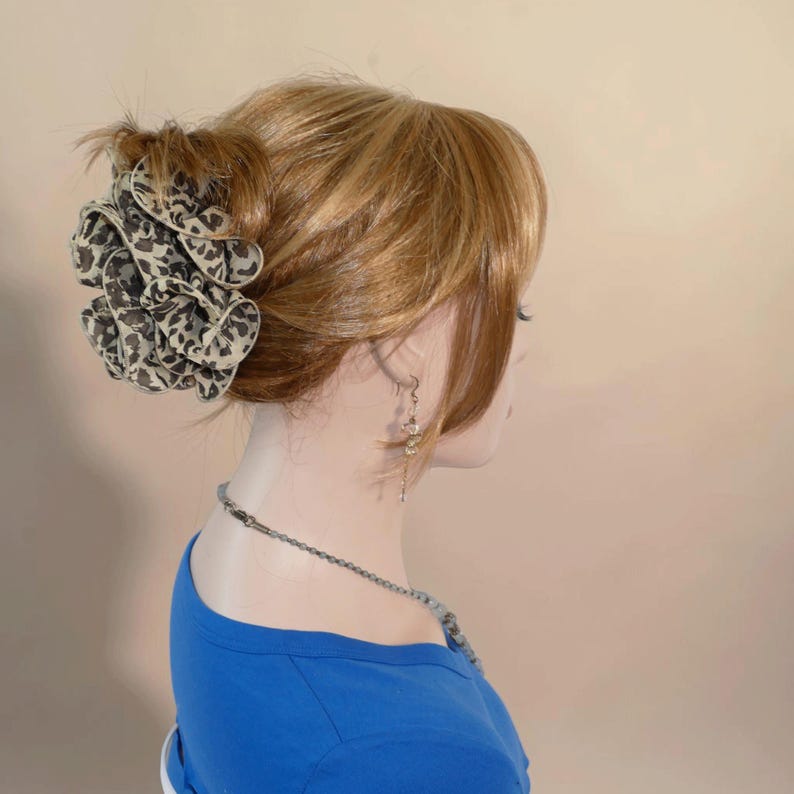 以下が含まれることがあります： 黒と白の動物柄の大きなフリルヘアアクセサリーが付いた、お団子ヘアスタイルのブロンドウィッグ。マネキンは青いトップスとシルバーのネックレスを着用しています。