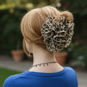 以下が含まれることがあります： ヒョウ柄の大きな重ね着ヘアアクセサリー。軽くて薄手の生地で作られており、髪に装着されています。背景はぼやけており、青いシャツとネックレスが見えます。