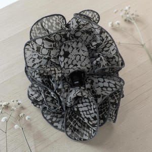 以下が含まれることがあります： グレーと黒の色合いの爬虫類プリント柄の布製フラワーヘアアクセサリー。花は複数のフリル層と暗い中心部を持っています。エッジは濃いグレーでトリミングされています。アクセサリーは明るい木製の表面にあります。