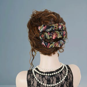 Puede incluir: Tres coleteros negros con un colorido estampado floral están apilados en el pelo de una mujer. Los coleteros presentan flores rojas, amarillas y azules sobre un fondo negro. La mujer lleva un top de encaje negro y un collar de perlas.