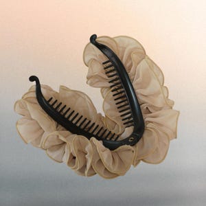 Op de afbeelding: Een zwarte bananen-haarklem van kunststof met een gebogen, getand ontwerp, versierd met lagen beige ruches van stof die een volumineus, decoratief effect creëren rondom de gehele rand van dit haaraccessoire.