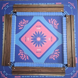 May include: A Mahjong game set with a blue and pink playing surface. The set includes wooden racks, blue tiles, and a central design with directional labels. The edges of the board have decorative floral patterns.