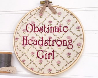 May include: A round embroidery hoop with a white background and pink floral pattern. The text "Obstinate Headstrong Girl" is embroidered in red.