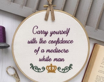 May include: An embroidery hoop with the text "Carry yourself with the confidence of a mediocre white man" in purple thread. A gold crown and green flourishes are embroidered below the text. A spool of purple thread and gold scissors are also visible.