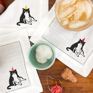 おもしろい刺繍入り猫カクテルナプキン：白いリネン100%に、パーティーハットをかぶった黒い太った猫が描かれています。猫好きの方への、新築祝いや誕生日のプレゼントにぴったりのユニークなギフトです。