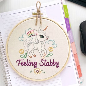 Finished embroidery hoop art on off white fabric with a colorful unicorn and the words 'feeling stabby' stitched in purple cursive.