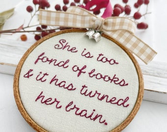 May include: A round, wooden-framed ornament with a white fabric background. The ornament features a burgundy embroidered quote: "She is too fond of books & it has turned her brain."