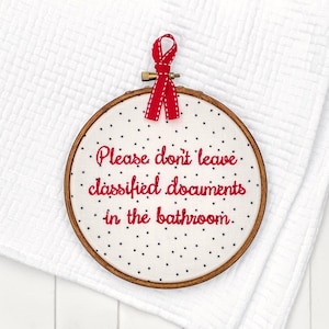 Peut inclure: Un cercle de broderie blanc avec un ruban rouge suspendu en haut. Le cercle a un fond blanc avec des étoiles noires et une broderie rouge qui dit "Veuillez ne pas laisser de documents classifiés dans la salle de bain."