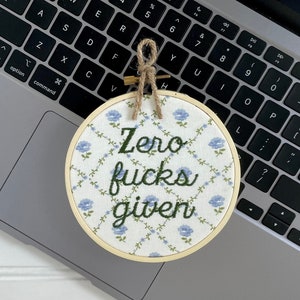 May include: A round embroidery hoop with the text "Zero fucks given" in green script. The fabric has a white background with a blue floral pattern. A jute twine loop is attached for hanging.