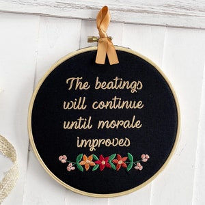 May include: A black embroidery hoop with the text "The beatings will continue until morale improves" in gold thread. The bottom features a floral design with orange, red, and pink flowers. A gold ribbon is attached for hanging.