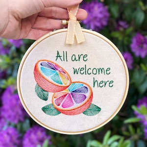 以下が含まれることがあります： 虹色の柑橘類のデザインと「All are welcome here」というテキストが刺繍されたフープアートです。