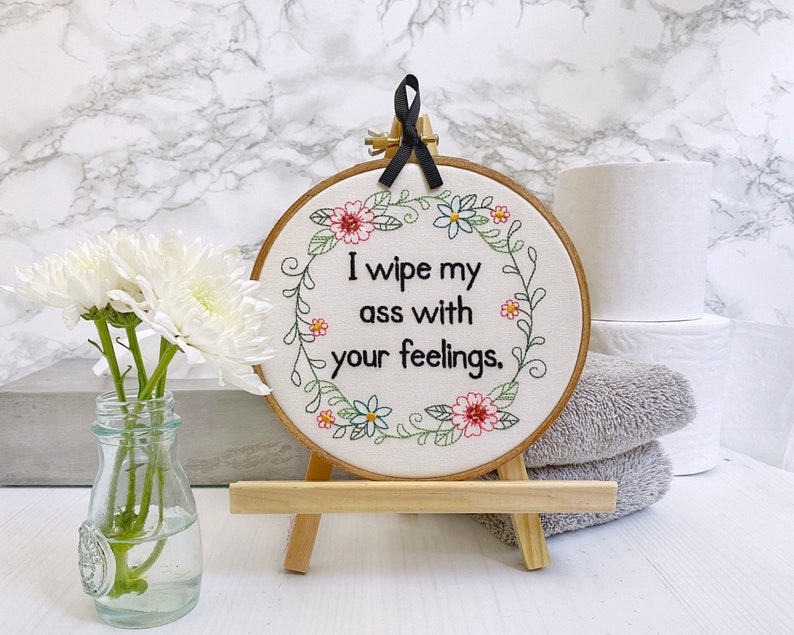 A floral finished embroidery art that has I wipe my ass with your feelings stitched in black thread in the center of a floral wreath. On off white woven cotton with a grosgrain ribbon for easy hanging. 6 inches in diameter total.