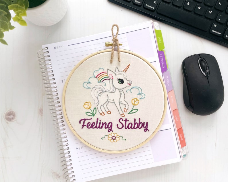 Finished embroidery hoop art on off white fabric with a colorful unicorn and the words 'feeling stabby' stitched in purple cursive.