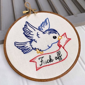 Könnte beinhalten: Stickerei auf einem Reifen mit einem blauen Vogel mit roten, weißen und blauen Akzenten, der ein Banner mit der Aufschrift "F*ck off" trägt.