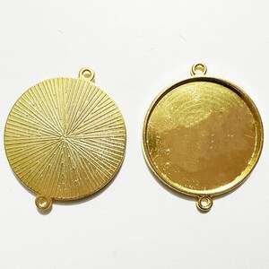 May include: Two gold-colored metal round pendant trays with a smooth surface and a textured surface. Each tray has a loop at the top for hanging.