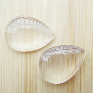 Op de afbeelding: Twee heldere glazen cabochons in druppelvorm met een rasterpatroon erin. De cabochons liggen op een licht houten oppervlak.