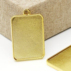 May include: Two gold-toned metal pendant trays with a rectangular shape and a loop at the top for hanging. The trays are blank and ready for resin or other embellishments.