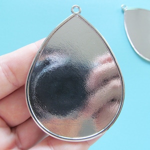 Könnte beinhalten: Silberner tropfenförmiger Anhänger mit einer glatten, reflektierenden Oberfläche. Der Anhänger hat oben eine Öse zum Anbringen einer Kette oder Schnur.