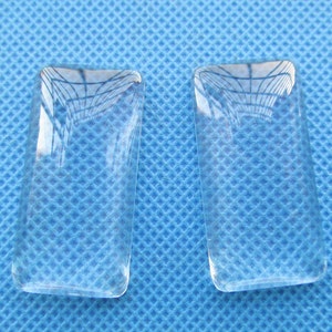 Op de afbeelding: Twee rechthoekige vormen van helder glas, mogelijk voor het maken van sieraden of knutselen, op een blauwe textuur achtergrond.