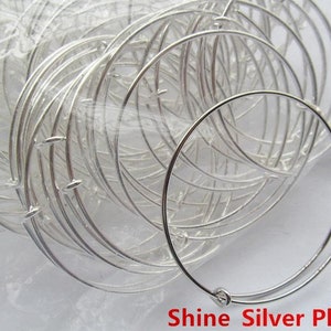 May include: A close-up of a pile of silver-plated bangle bracelets. The bracelets are all the same size and are made of thin metal. The bracelets are arranged in a pile, with some of the bracelets overlapping each other. The bracelets are shiny and reflect the light. The text "Shine Silver Plated" is visible at the bottom of the image.