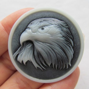 Könnte beinhalten: Ein weißer Kamee mit einer detaillierten Schnitzerei eines Adlerkopfes. Der Adler blickt nach links und hat einen scharfen Schnabel und durchdringende Augen. Der Kamee ist vor einem schwarzen Hintergrund platziert.